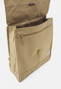 Tan fabric messenger bag with a zippered compartment, flap closure, and front pocket. Features a logo tag and reinforced stitching.