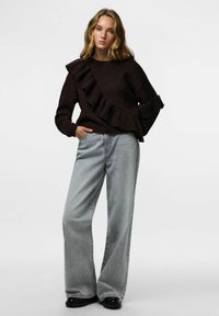 Brązowy dzianinowy sweter z falbankami po jednej stronie, zestawiony z jasnoszarymi szerokimi dżinsami i czarnymi lakierowanymi butami.