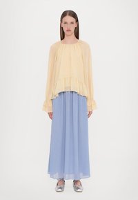 Lichtgele, semi-doorzichtige blouse met een gerimpelde zoom, gecombineerd met een lange, vloeiende blauwe rok. Zilveren ballerina's maken de outfit compleet.