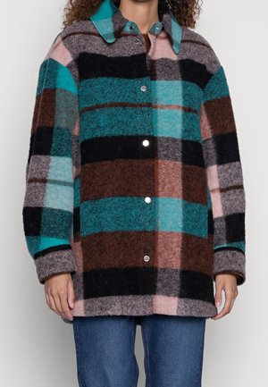 Person, die eine zugeknöpfte, übergroße karierte Jacke in Türkis, Braun, Schwarz, Pink und Grau trägt, kombiniert mit blauen Jeans.
