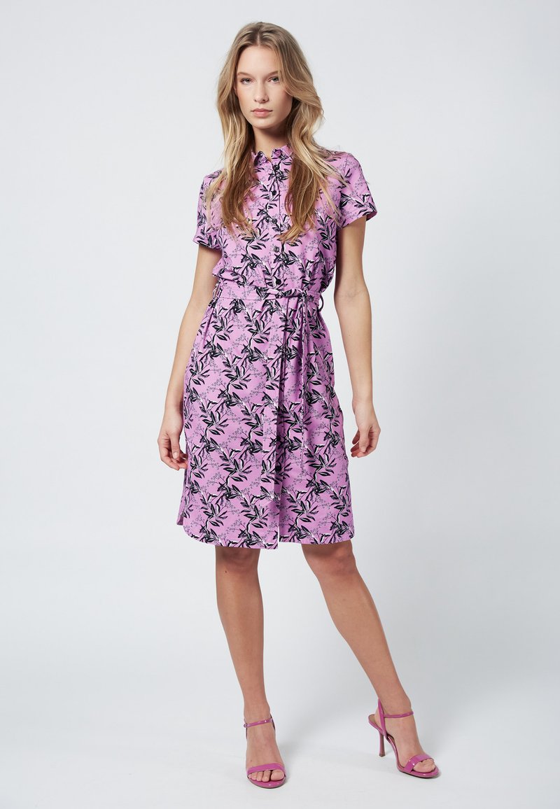 Abito floreale rosa con maniche corte, motivo a foglie nere e vita cinchata. Accompagnato da tacchi a strappo rosa coordinati.