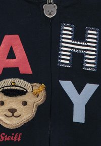 Námořnický zip-up s vyšitými červenými a modrými písmeny "A", "H" a "Y". Obsahuje aplikaci s obličejem plyšového medvěda s námořnickou čepicí a zlatým akcentem.