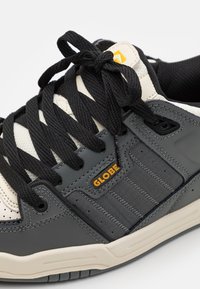 Gray and cream sneaker with black laces, rubber sole, and mesh detailing. Features a logo and textured accents on the side and vamp.