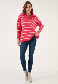 Rožinė ir balta juostuota zip-up megztinis su apykakle, derintas su tamsiai mėlynomis aptemptomis džinsomis ir smėlio spalvos kulkšninių botų ant vientiso fono.