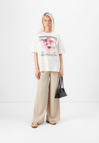 Beige grafisk t-shirt med texten "HOPLESS ROMANTIC", tillsammans med lösa, strukturerade ljusbeige byxor och svart handväska. Skorna är mönstrade.