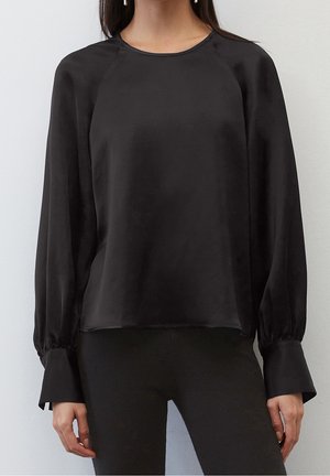 Kvinde iført en sort satinbluse med lange ærmer, brede manchetter og rund halsudskæring, kombineret med sorte bukser.