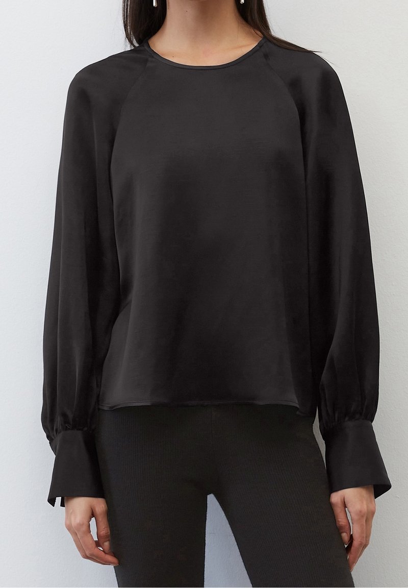 Femme portant un chemisier en satin noir à manches longues avec de larges poignets et un col rond, associé à un pantalon noir.