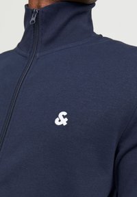 Mornarska jakna na zip s visokim ovratnikom, izrađena od glatke tkanine. Ima bijeli izvezeni logo na lijevoj strani prsa i odgovarajući patentni zatvarač.