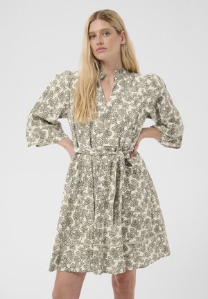 Femme aux longs cheveux blonds portant une robe fleurie mi-longue aux manches bouffantes et ceinturée à la taille, debout les mains sur les hanches.