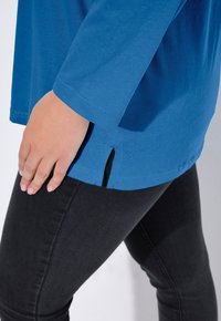 Blaues Langarmhemd mit seitlichem Schlitz, kombiniert mit schwarzen, eng anliegenden Jeans. Der Stoff wirkt weich und glatt, mit wenigen Nähten.