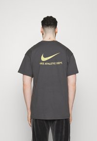 Šedé bavlněné tričko s žlutým logem Nike a textem "NIKE ATHLETIC DEPT." na zádech. Krátké rukávy a kulatý výstřih.