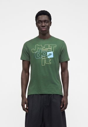 Jeune homme portant un t-shirt vert Nike avec le slogan "Just do it" et un pantalon noir, debout devant un fond clair uni.
