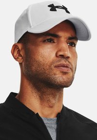 Hvid kasket med et sort Under Armour-logo, med en struktureret design og buet skygge, båret med en sort jakke over en grå skjorte.