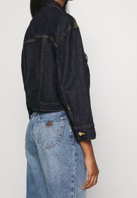 Krátká džínová bunda tmavě modré barvy s kontrastním žlutým šitím, knoflíkovými manžetami a náprsními kapsami, doplněná o světle modré džíny s vysokým pasem.