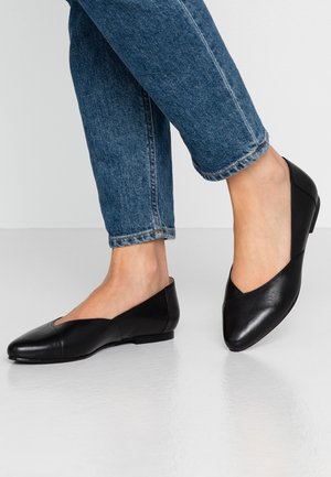 Personne portant un jean bleu slim et des chaussures plates en cuir noir debout sur un sol clair.