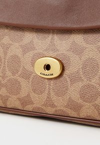 Borsa a mano marrone con motivo, caratterizzata da uno sfondo di un beige chiaro, tessuto testurizzato e una chiusura ovale dorata con la dicitura "COACH" incisa.
