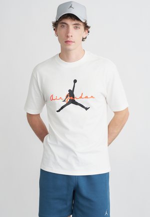 Young man wearing white Air Jordan t-shirt, grey Air Jordan cap, and blue shorts standing against a plain white background.