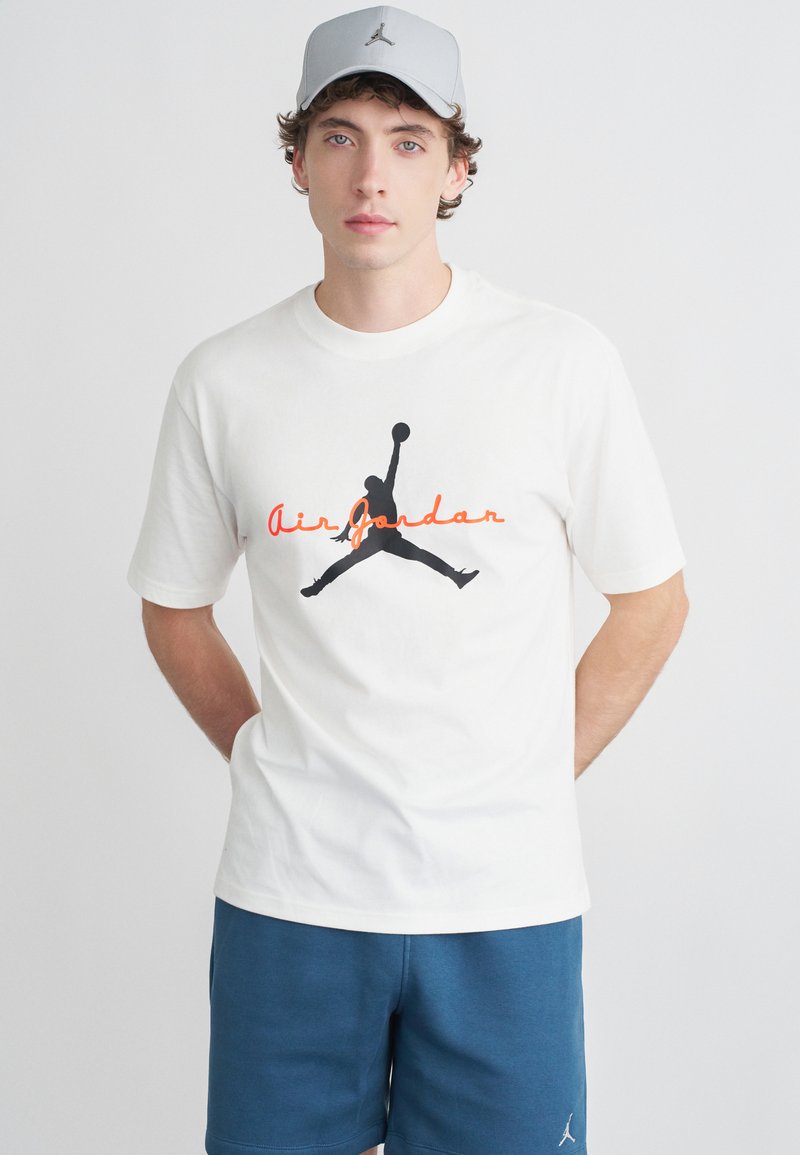 Νεαρός άνδρας που φοράει λευκό μπλουζάκι Air Jordan, γκρι καπέλο Air Jordan και μπλε σορτς, στέκεται μπροστά σε απλό λευκό φόντο.