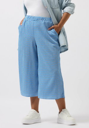 Person wearing blue and white checkered wide-leg cropped pants, white sneakers, white top, and light denim jacket with hands in pockets.