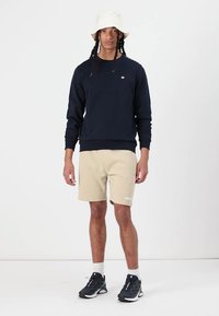 Marine sweatshirt, beige shorts og sorte sneakers med hvide detaljer. Modellen bærer en hvid bucket hat, hvilket fremhæver et afslappet outfit.