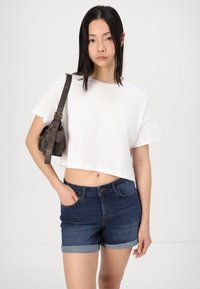 T-shirt branca cropped em tecido suave, acompanhada de calções de ganga azul-escuro com barras dobradas, apresentando passantes para cinto e bolsos frontais.