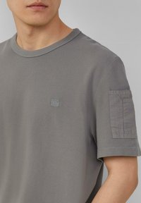 Grijze katoenen t-shirt met een ronde hals, korte mouwen en een gestructureerde zak op de rechtermouw met gestikte details.