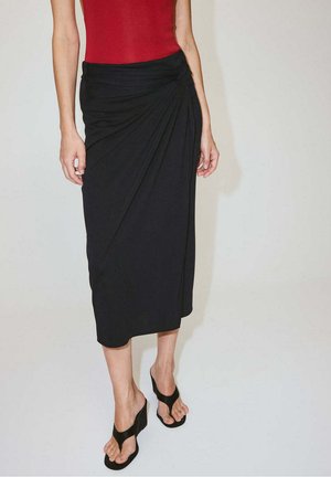 Femme portant une jupe midi noire avec un détail de nœud sur le côté, un haut rouge sans manches et des sandales compensées noires avec des brides aux orteils.