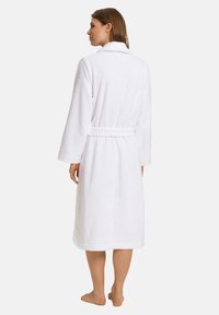 Vit frottébadrock, knälång, med långa ärmar, midja med bälte och sjalkrage. Mjuk struktur med ett enfärgat mönster.