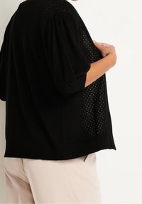 Osoba w czarnej, dziurkowanej, dzianinowej bluzce z bufkami oraz jasnobeżowych spodniach, trzymająca przód bluzki.