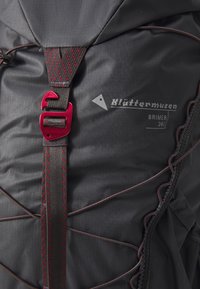 Schwarzer Rucksack mit strukturiertem Stoff, roten Akzenten, einem auffälligen Logo und einem einzigartigen Design von Frontgurt und Schnalle. Kapazität von 24 Litern.