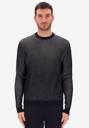 Man with short dark hair and beard wearing a black and gray vertically striped long-sleeve sweater, standing against a plain light background.