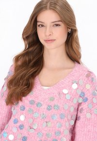 Różowy sweter z dzianiny z dekoltem w serek, ozdobiony iryzującymi, okrągłymi cekinami w różnych rozmiarach na przodzie. Teksturowana tkanina.