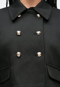 Čierny dvojradový kabát z textúrovanej látky, s veľkým golierom a detailmi zlatých gombíkov, s dvoma bočnými vreckami.