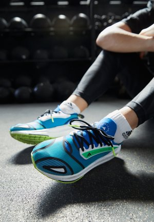 Des chaussures de sport bleues avec des accents verts, présentant une tige texturée, des lacets noirs et une semelle robuste. Des chaussettes blanches avec un logo sont visibles.