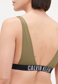 Maslinasto zeleni sportski grudnjak sa širokim naramenicama i odsječenim leđima. Sadrži crni pojas s natpisom "CALVIN KLEIN" u bijelom slovima. Glatka tekstura.