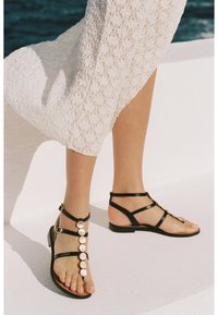 Sandali neri piatti con molte cinghie sottili e dettagli metallici rotondi. Le cinghie sono lisce e la suola è piatta e marrone.