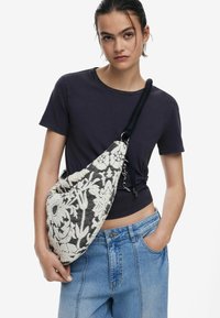 Strukturierte Stofftasche mit schwarzem und cremefarbenem Blumenmuster, lässigem Design und einem schwarz gestreiften Griff. Im Crossbody-Stil getragen.