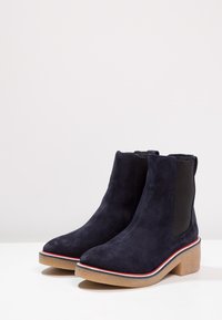 Botines de ante azul marino con punta afilada, paneles laterales elásticos y un tacón cuadrado texturizado beige, que presenta un borde contrastante en rojo y azul.