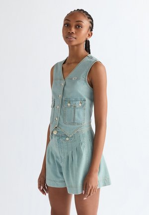 Jonge vrouw met gevlochten haar, gekleed in een lichtblauw mouwloos spijkervest en bijpassende shorts met hoge taille, staand tegen een effen witte achtergrond.