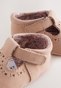 Chaussures bébés beige douces avec perforations en forme de cœur, doublure en tissu floral et bandes velcro ajustables, montrées en gros plan sur fond neutre.