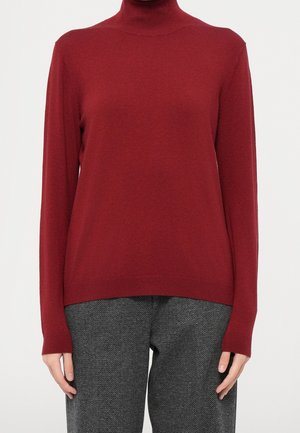 Person wearing a red long-sleeve turtleneck sweater and gray herringbone patterned pants with hands relaxed by sides.