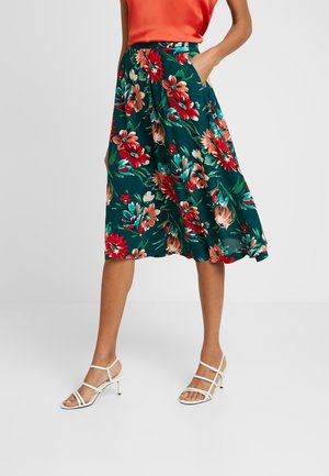 Kvinde iført en teal midikjole med blomster i røde og pink farver, en orange top og hvide sandaler med hæl og stropper.