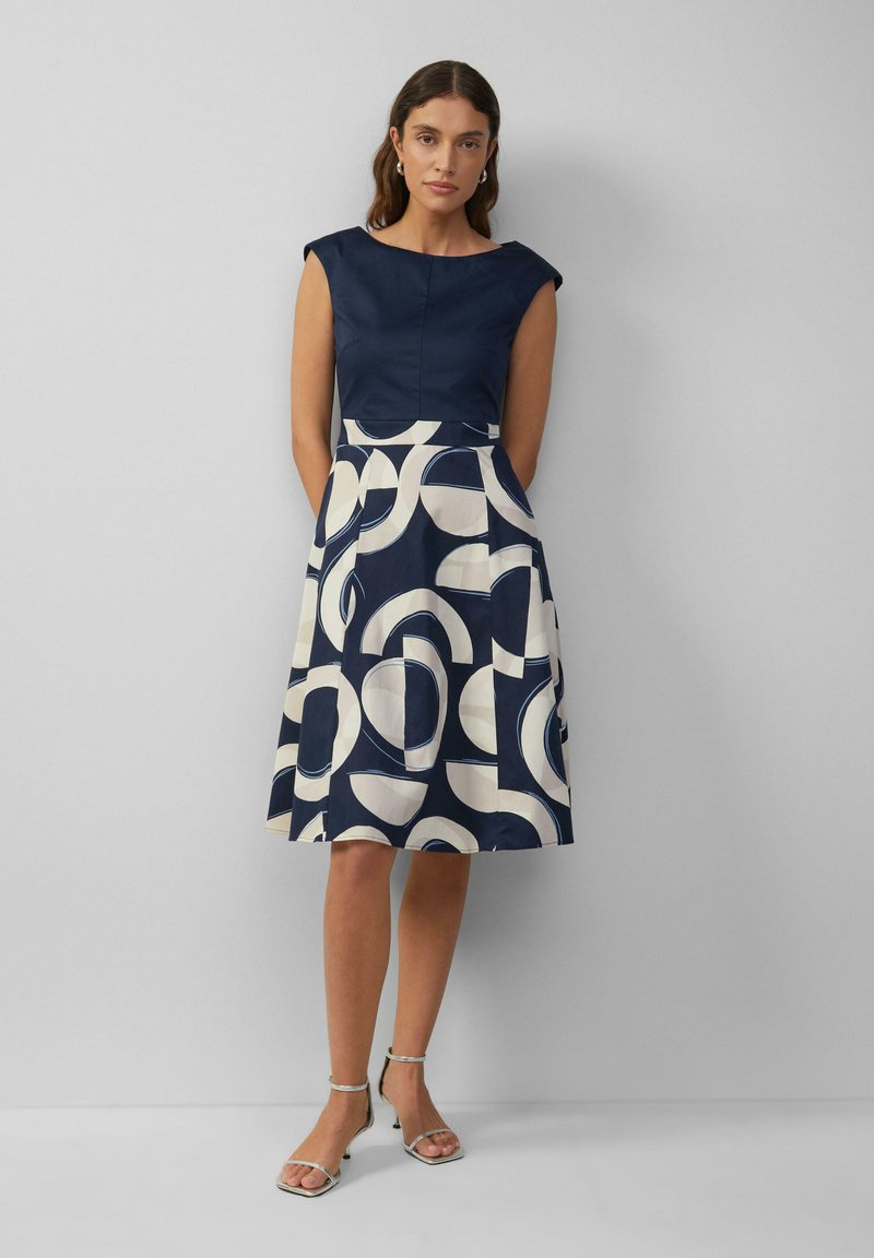 Navyblaues Kleid mit tailliertem Oberteil und A-Linie-Rock, der ein auffälliges cremefarbenes abstraktes Muster zeigt. Glattes Material und Kapellärmeln.