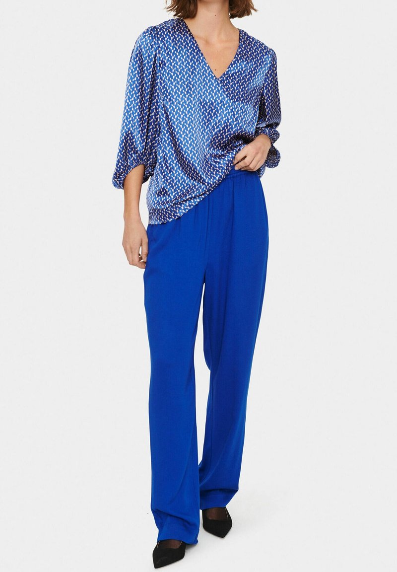 Una donna indossa una blusa blu a fantasia e pantaloni larghi blu, accessoriate con scarpe nere, in piedi contro uno sfondo bianco.