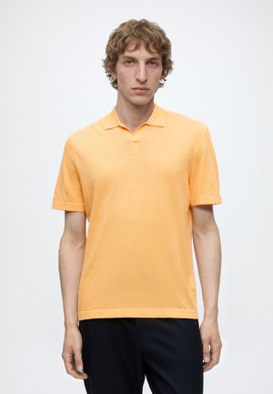 Jeune homme aux cheveux bouclés portant un polo à manches courtes couleur pêche et un pantalon foncé, debout devant un fond blanc uni.