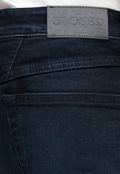 Jean en denim bleu foncé avec des coutures visibles sur les poches arrière et un patch en cuir gris avec la marque "CLOSED" sur la ceinture.