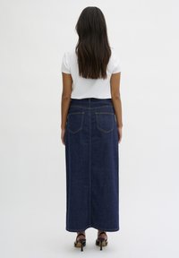 Gonna maxi in denim di colore blu scuro, con una vestibilità dritta, cuciture a vista, due tasche posteriori e una vita alta. Indossata con una maglietta bianca e tacchi.