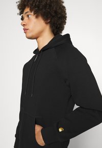 Schwarze Kapuzenjacke aus Baumwolle, mit einer Vorderjacke und goldfarbener Logo-Akzent auf dem Ärmel. Glatte Textur und lockere Passform.