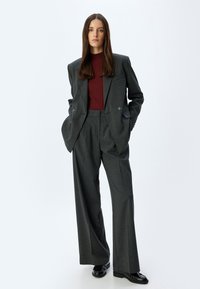 Grijze op maat gemaakte kostuum met wijde broekspijpen en een dubbelgeknoopte blazer, gedragen over een bordeauxrode geribde top. Zwarte schoenen maken de outfit af.