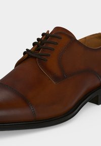 Chaussure habillée pour homme en cuir marron avec lacets noirs, coutures sur le bout et petit talon noir sur fond blanc.